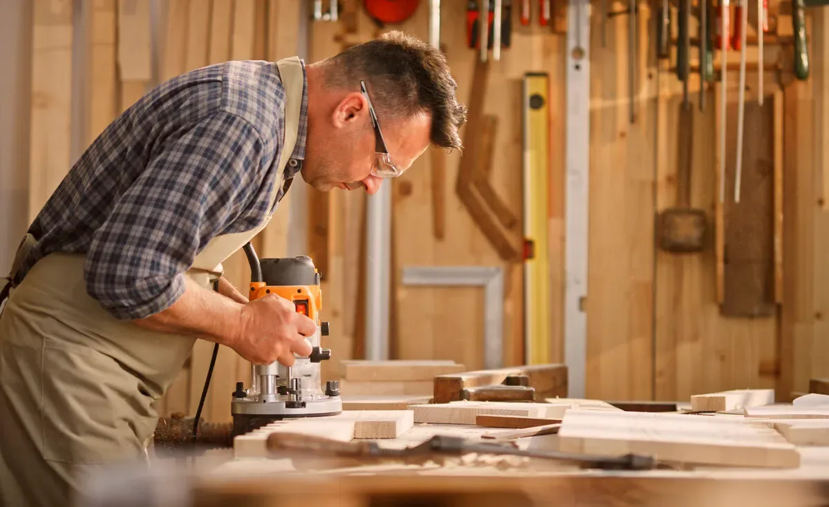 [An overhead shot of a woodworker using a router to create a decorative edge on a walnut tabletop, with wood shavings curling away from the bit]